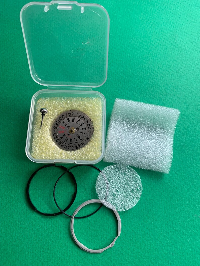 May include: A watch repair kit with various components. The kit includes a silver-colored watch dial with day and date indicators, a small screw, black rubber rings, and a clear plastic case. The items are on a green surface.
