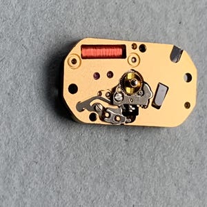 May include: Close-up of a watch movement with a gold-colored base. The movement features a red coil, small gears, and various silver-colored components. Several small holes and dark accents are visible on the surface.