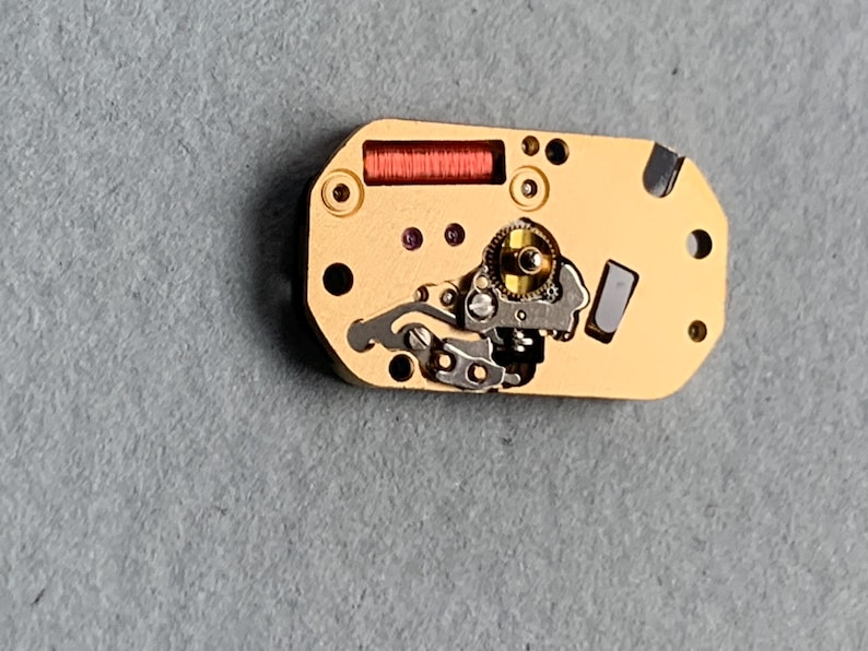 May include: Close-up of a watch movement with a gold-colored base. The movement features a red coil, small gears, and various silver-colored components. Several small holes and dark accents are visible on the surface.