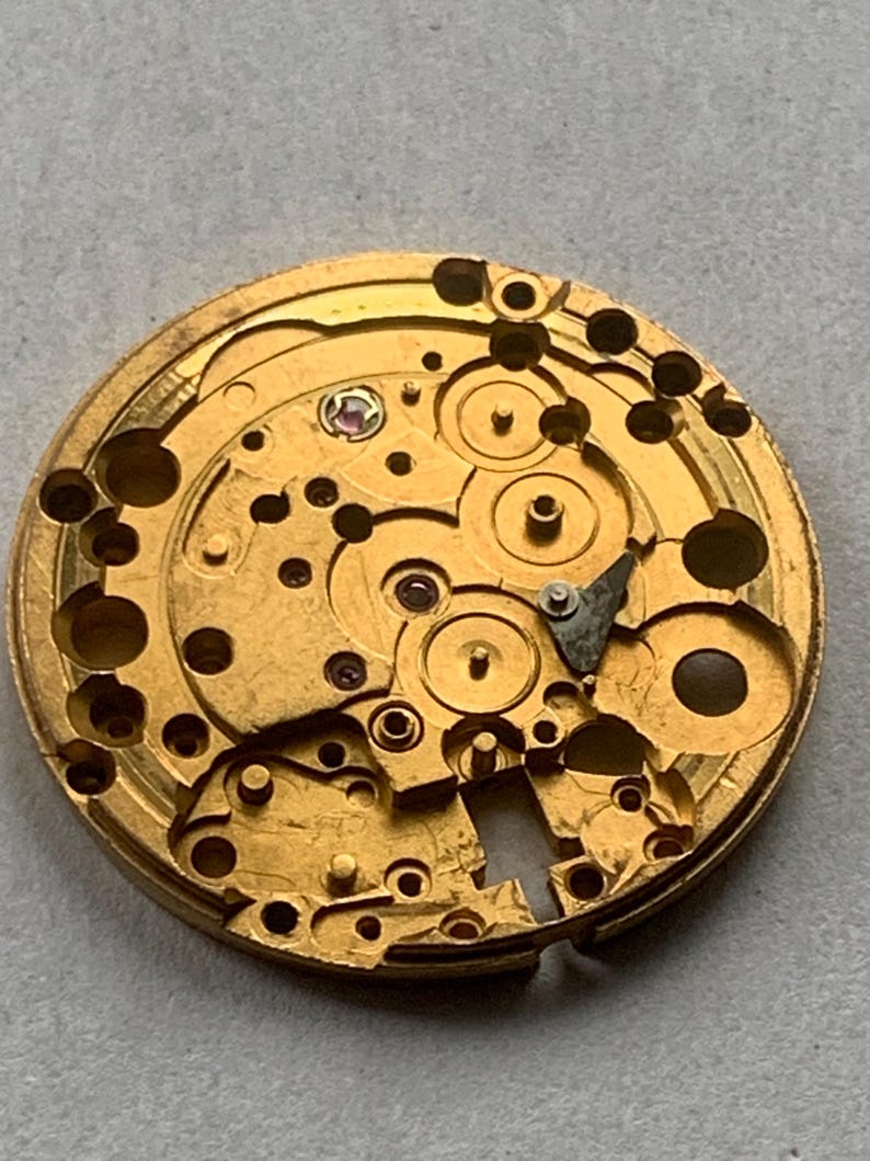 May include: Close-up of a gold-colored watch movement with intricate gears, springs, and jewels. The mechanical components are arranged in a circular pattern, showcasing the precision of the watch mechanism.
