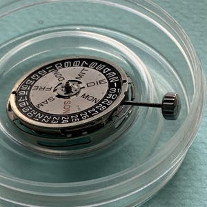 May include: A watch movement with a silver-colored dial and black markings, including the days of the week and numbers 1-31. The movement is housed in a clear plastic container. A small, black crown is attached.