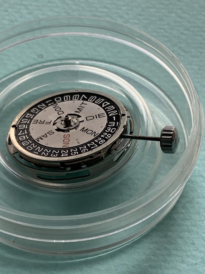 May include: A watch movement with a silver-colored dial and black markings, including the days of the week and numbers 1-31. The movement is housed in a clear plastic container. A small, black crown is attached.