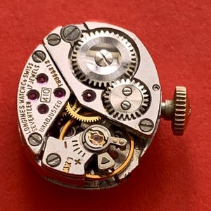 May include: Close-up of a vintage Longines watch movement with intricate gears, jewels, and a winding stem. The watch mechanism is silver-toned with gold accents and the text "Longines Watch Co Swiss 17 Jewels" is visible.