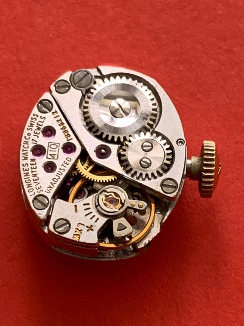 May include: Close-up of a vintage Longines watch movement with intricate gears, jewels, and a winding stem. The watch mechanism is silver-toned with gold accents and the text "Longines Watch Co Swiss 17 Jewels" is visible.