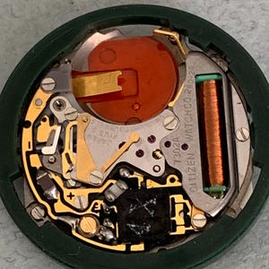 May include: Close-up of a Citizen watch movement, showcasing intricate internal components. The watch mechanism features gold-colored gears, a copper coil, and a large orange battery. The movement is housed in a dark green circular frame.