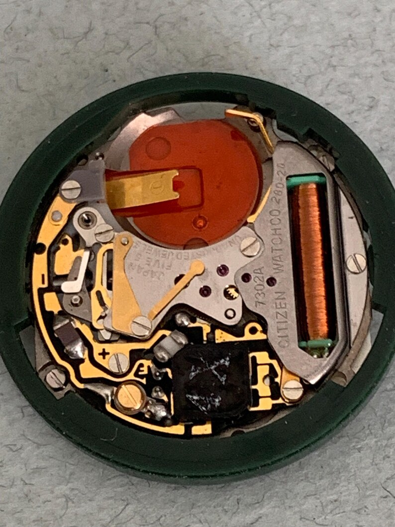 May include: Close-up of a Citizen watch movement, showcasing intricate internal components. The watch mechanism features gold-colored gears, a copper coil, and a large orange battery. The movement is housed in a dark green circular frame.