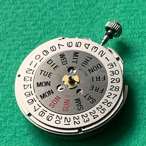 May include: A silver watch movement with a day and date display. The dial features numbers 1-31, and days of the week in English, with a small crown. The background is a textured green surface.
