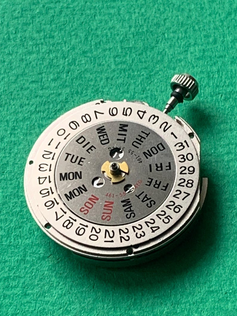 May include: A silver watch movement with a day and date display. The dial features numbers 1-31, and days of the week in English, with a small crown. The background is a textured green surface.