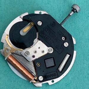 May include: A close-up of an Orient watch movement, model 56910 2j. The watch mechanism features a black plate with the Orient logo and "Japan" text. It has silver, gold, and copper components, including a winding stem and a coil.