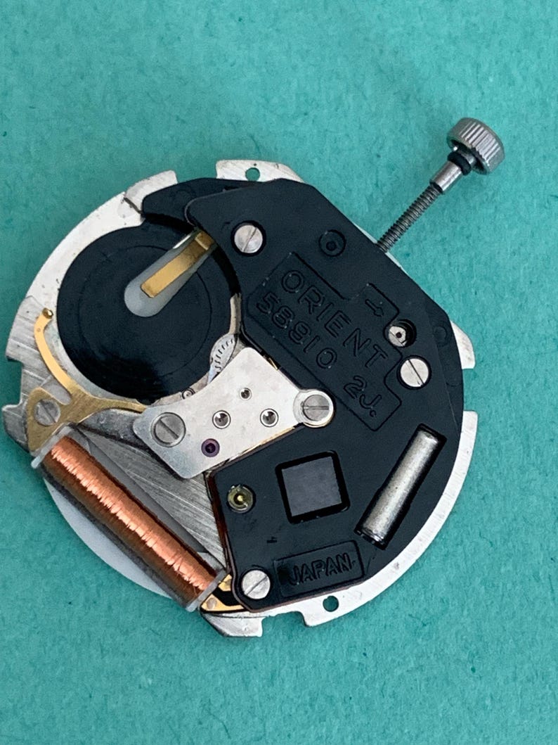 May include: A close-up of an Orient watch movement, model 56910 2j. The watch mechanism features a black plate with the Orient logo and "Japan" text. It has silver, gold, and copper components, including a winding stem and a coil.
