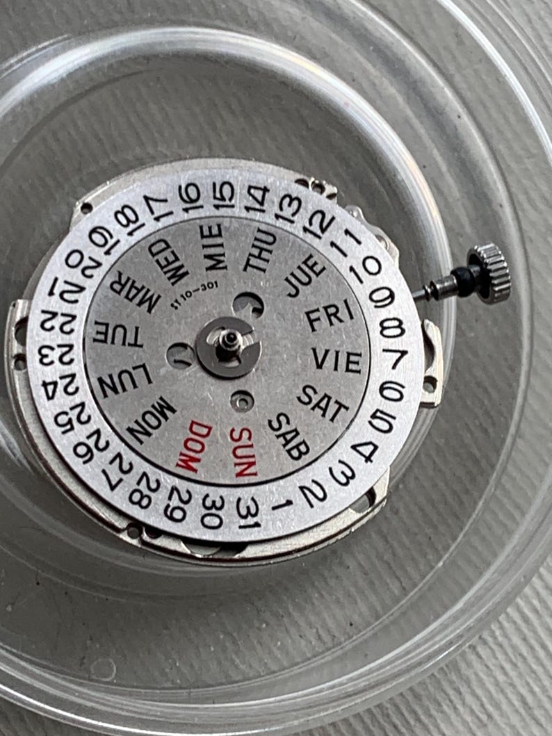 Pu&ograve; includere: Un movimento di orologio argentato con un quadrante del calendario che mostra i giorni della settimana in pi&ugrave; lingue. Il quadrante presenta i numeri da 1 a 31 e i giorni della settimana. Sono visibili una piccola corona e uno stelo. Il movimento &egrave; in un contenitore di plastica trasparente.