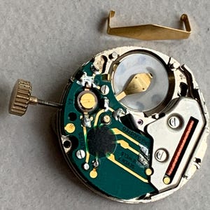 Puede incluir: Primer plano de un movimiento de reloj con una placa de circuito verde, componentes dorados y una corona dorada. Una pieza met&aacute;lica dorada separada est&aacute; encima del movimiento. El movimiento del reloj tiene el texto "SWISS" y "SEVEN 7 JEWELS".