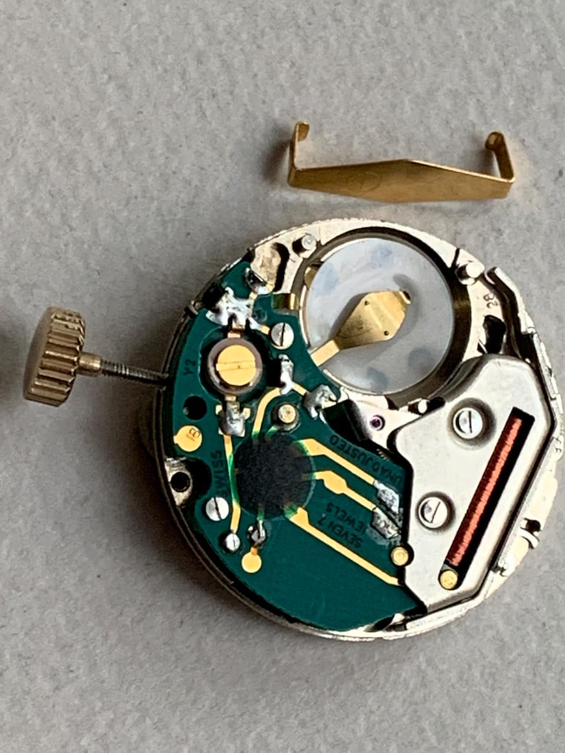 Puede incluir: Primer plano de un movimiento de reloj con una placa de circuito verde, componentes dorados y una corona dorada. Una pieza met&aacute;lica dorada separada est&aacute; encima del movimiento. El movimiento del reloj tiene el texto "SWISS" y "SEVEN 7 JEWELS".