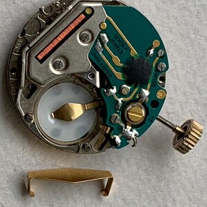 Puede incluir: Primer plano de un movimiento de reloj con una placa de circuito verde, componentes dorados y una peque&ntilde;a corona dorada. Las palabras "SEVEN 7 JEWELS" y "UNADJUSTED" son visibles. Un cierre de metal dorado en la parte inferior.