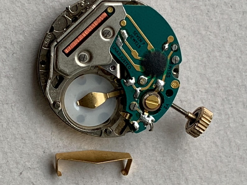 Puede incluir: Primer plano de un movimiento de reloj con una placa de circuito verde, componentes dorados y una peque&ntilde;a corona dorada. Las palabras "SEVEN 7 JEWELS" y "UNADJUSTED" son visibles. Un cierre de metal dorado en la parte inferior.
