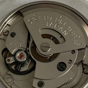May include: Close-up of a watch movement with the text "ORIENT 21J JAPAN 46943" visible. The mechanism features various gears, screws, and a jewel-toned ruby. The overall color scheme is silver and metallic.