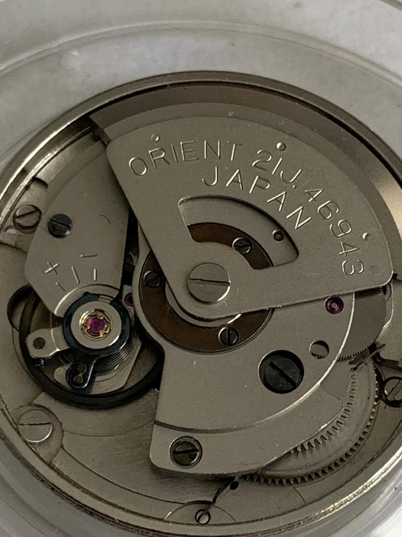 May include: Close-up of a watch movement with the text "ORIENT 21J JAPAN 46943" visible. The mechanism features various gears, screws, and a jewel-toned ruby. The overall color scheme is silver and metallic.