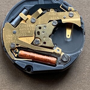 May include: Close-up of a Citizen watch movement. The watch mechanism features gold-toned metal components, a copper-colored coil, and the text "CITIZEN WATCH CO. 3801A UNADJUSTED ONE 1 JEWEL" and "PUSH 260-31 JAPAN". The movement is set in a gray plastic casing.
