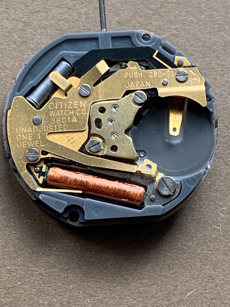 May include: Close-up of a Citizen watch movement. The watch mechanism features gold-toned metal components, a copper-colored coil, and the text "CITIZEN WATCH CO. 3801A UNADJUSTED ONE 1 JEWEL" and "PUSH 260-31 JAPAN". The movement is set in a gray plastic casing.