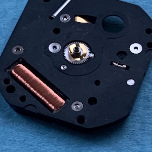 May include: Close-up of a black watch movement with a copper coil, small gears, and silver components. The watch mechanism is set against a blue background. The intricate details of the watch parts are visible.