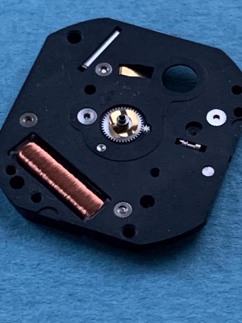 May include: Close-up of a black watch movement with a copper coil, small gears, and silver components. The watch mechanism is set against a blue background. The intricate details of the watch parts are visible.