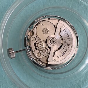 May include: A close-up of a watch movement, featuring intricate gears and components. The watch mechanism is encased in a clear, circular container. The text "CITIZEN 21 JEWELS JAPAN" is visible on the watch face.