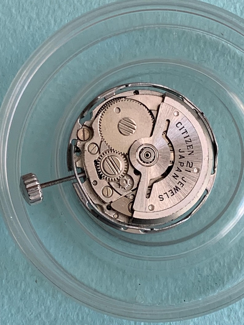 May include: A close-up of a watch movement, featuring intricate gears and components. The watch mechanism is encased in a clear, circular container. The text "CITIZEN 21 JEWELS JAPAN" is visible on the watch face.