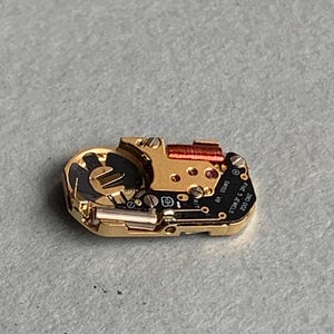 May include: A close-up of a watch movement with gold-colored metal components, including screws, gears, and a red coil. The movement has a black circuit board with the text "SWISS V8 FIVE 5 JEWELS 280 002".