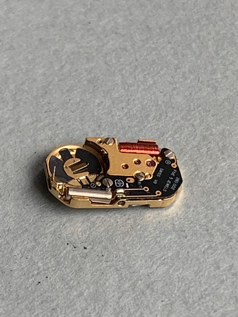 May include: A close-up of a watch movement with gold-colored metal components, including screws, gears, and a red coil. The movement has a black circuit board with the text "SWISS V8 FIVE 5 JEWELS 280 002".