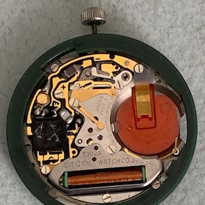 May include: A close-up of a Citizen watch movement, showcasing intricate internal components. The watch mechanism features gold-colored gears, a red battery, and the text "JAPAN FIVE 5 JEWELS". The watch's green casing surrounds the internal parts.