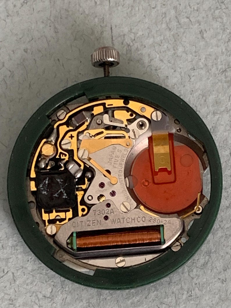 May include: A close-up of a Citizen watch movement, showcasing intricate internal components. The watch mechanism features gold-colored gears, a red battery, and the text "JAPAN FIVE 5 JEWELS". The watch's green casing surrounds the internal parts.