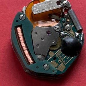 May include: Close-up of a watch movement with intricate gears, circuits, and metal components. The movement features a green circuit board, copper coils, and a small, black component. The text "SWISS UNADJUSTED" is visible.