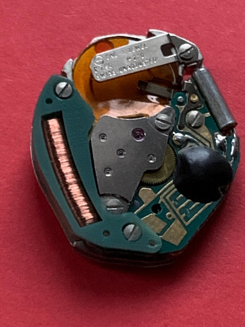May include: Close-up of a watch movement with intricate gears, circuits, and metal components. The movement features a green circuit board, copper coils, and a small, black component. The text "SWISS UNADJUSTED" is visible.