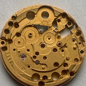 May include: Close-up of a gold-colored watch movement. The intricate mechanism features numerous gears, holes, and small components. A small, red jewel is visible. The watch movement is a complex piece of machinery.