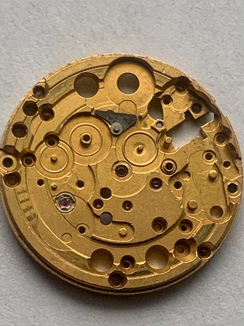 May include: Close-up of a gold-colored watch movement. The intricate mechanism features numerous gears, holes, and small components. A small, red jewel is visible. The watch movement is a complex piece of machinery.