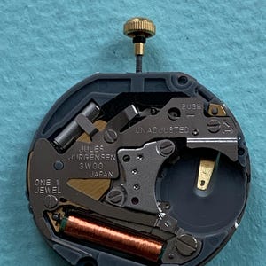 K&ouml;nnte beinhalten: Nahaufnahme eines Uhrwerks mit der Aufschrift "Jules Jurgensen 3W00 Japan" und "One 1 Jewel". Das Uhrwerk hat eine goldfarbene Krone, eine kupferfarbene Spule und verschiedene silber- und goldfarbene Komponenten. Der Hintergrund ist hellblau.