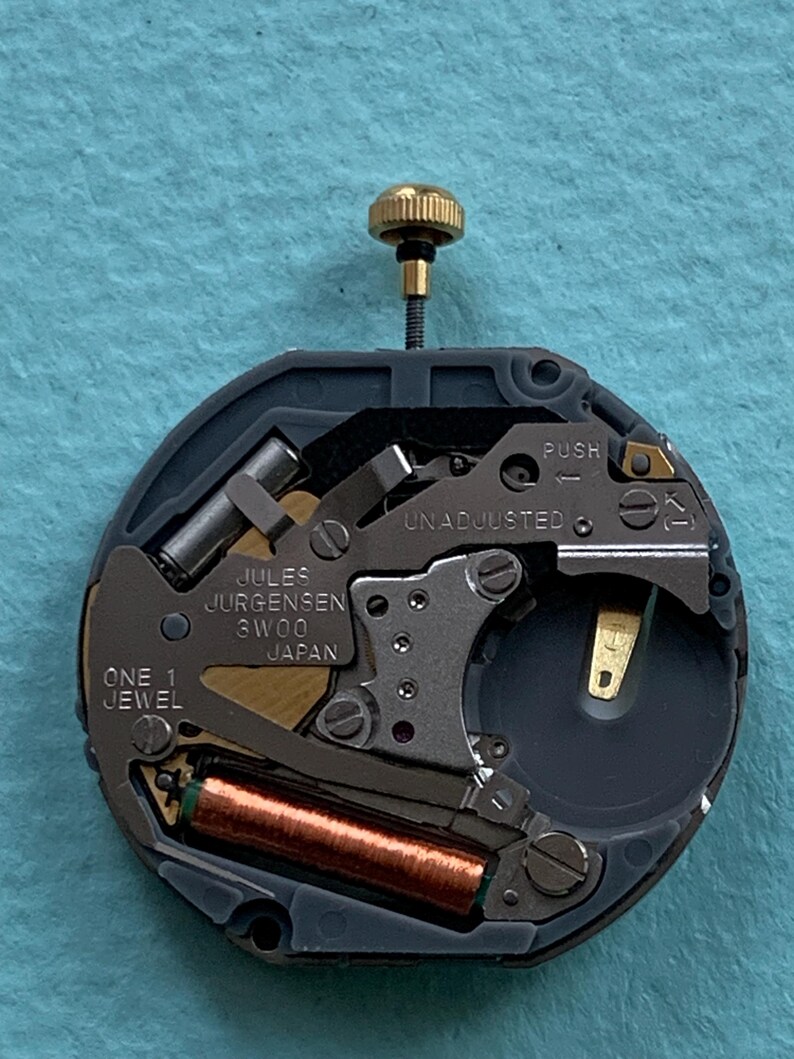 K&ouml;nnte beinhalten: Nahaufnahme eines Uhrwerks mit der Aufschrift "Jules Jurgensen 3W00 Japan" und "One 1 Jewel". Das Uhrwerk hat eine goldfarbene Krone, eine kupferfarbene Spule und verschiedene silber- und goldfarbene Komponenten. Der Hintergrund ist hellblau.
