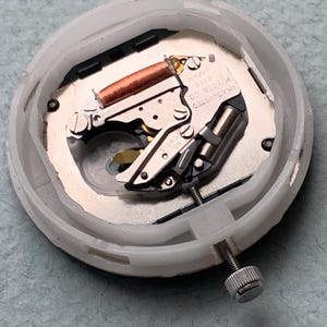 Puede incluir: Un movimiento de reloj con una carcasa de pl&aacute;stico blanco. Los componentes internos incluyen una bobina de cobre, piezas met&aacute;licas de color plateado y un tornillo. El texto "MIYOTA CO. JAPAN 2115" es visible en una placa met&aacute;lica.