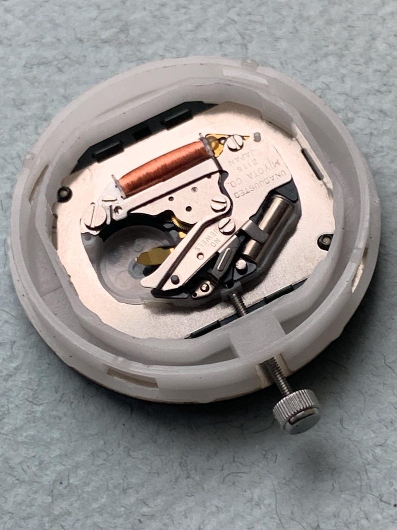 Puede incluir: Un movimiento de reloj con una carcasa de pl&aacute;stico blanco. Los componentes internos incluyen una bobina de cobre, piezas met&aacute;licas de color plateado y un tornillo. El texto "MIYOTA CO. JAPAN 2115" es visible en una placa met&aacute;lica.