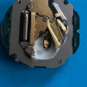 May include: Detailed view of a watch movement, revealing its complex internal mechanisms. The image features gold-coloured gears, a dark cylindrical coil, and a silver frame. The watch movement is set against a blue backdrop.