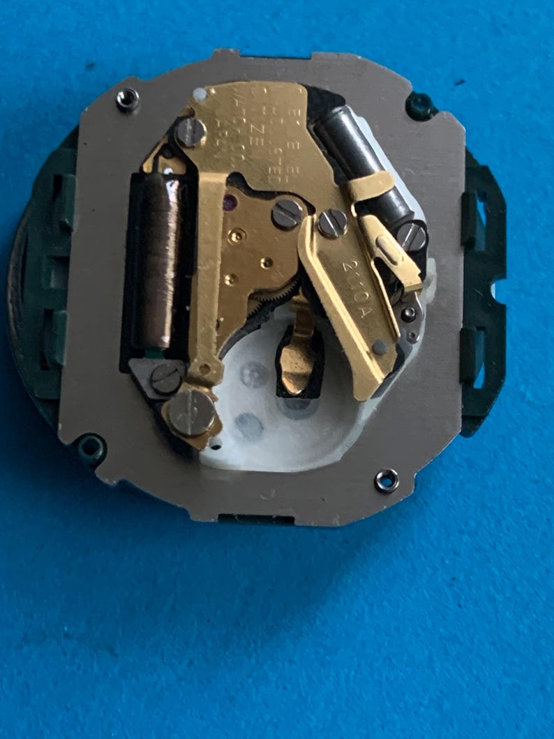 May include: Detailed view of a watch movement, revealing its complex internal mechanisms. The image features gold-coloured gears, a dark cylindrical coil, and a silver frame. The watch movement is set against a blue backdrop.