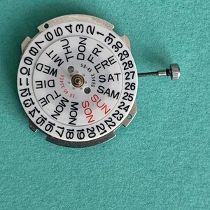 May include: A watch movement with a white dial showing the days of the week and numbers 1-31. It has a central mechanism and a small screw. The background is teal.