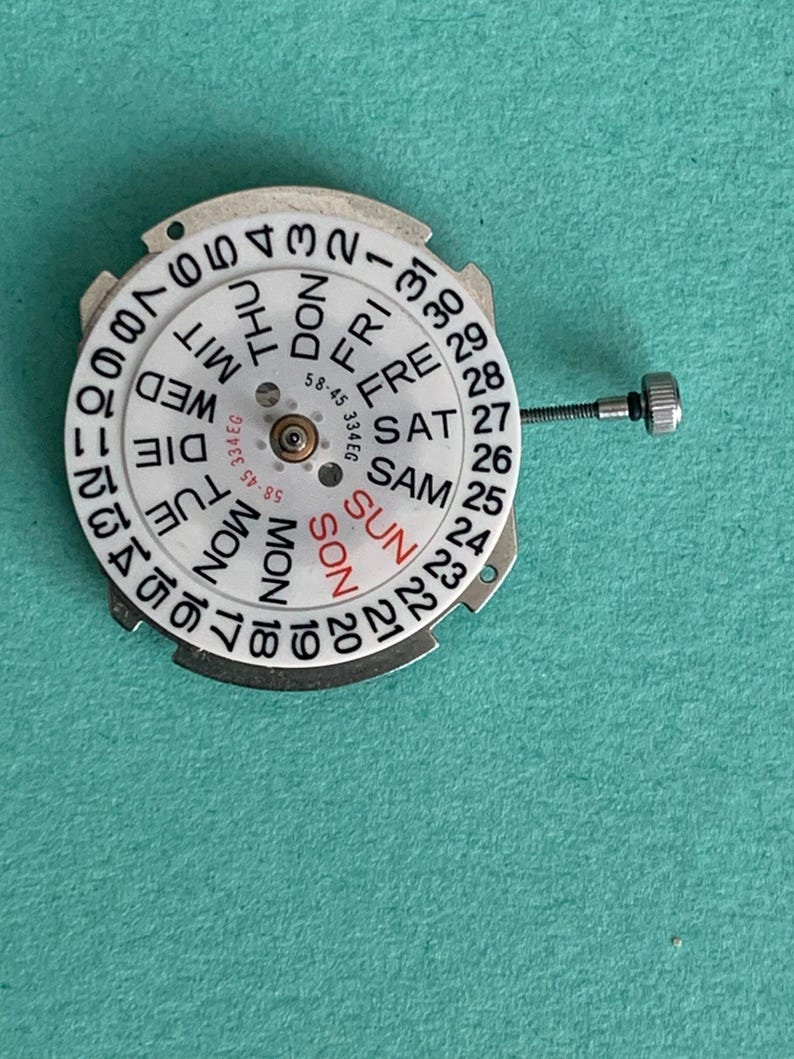 May include: A watch movement with a white dial showing the days of the week and numbers 1-31. It has a central mechanism and a small screw. The background is teal.