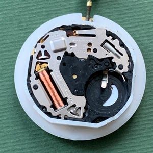 K&ouml;nnte beinhalten: Nahaufnahme eines Uhrwerks mit wei&szlig;er, kreisf&ouml;rmiger Basis. Die inneren Komponenten umfassen eine Kupferspule, silberfarbene Metallplatten und ein schwarzes Kunststoffteil. Eine silberfarbene Krone ragt oben heraus. Der Hintergrund ist einfarbig gr&uuml;n.