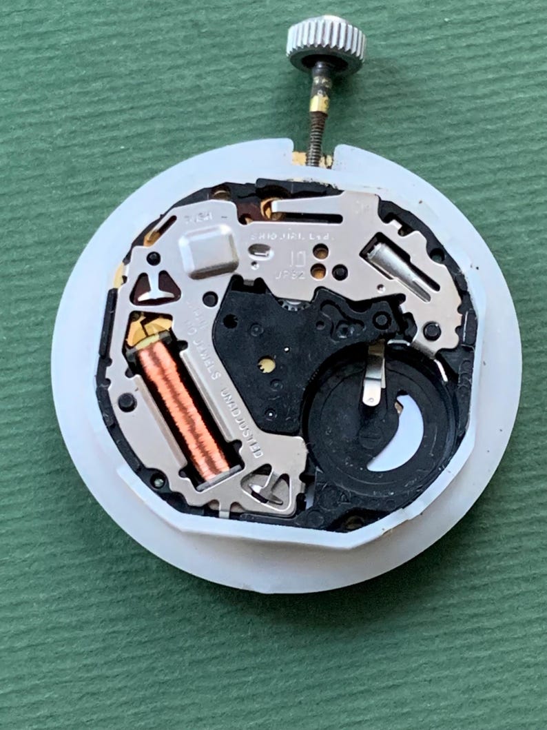 K&ouml;nnte beinhalten: Nahaufnahme eines Uhrwerks mit wei&szlig;er, kreisf&ouml;rmiger Basis. Die inneren Komponenten umfassen eine Kupferspule, silberfarbene Metallplatten und ein schwarzes Kunststoffteil. Eine silberfarbene Krone ragt oben heraus. Der Hintergrund ist einfarbig gr&uuml;n.