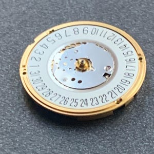 May include: A gold-toned watch movement with a white dial featuring black numerals from 1 to 31, indicating the days of the month. The central mechanism has a small gold-colored post. The watch component is circular.