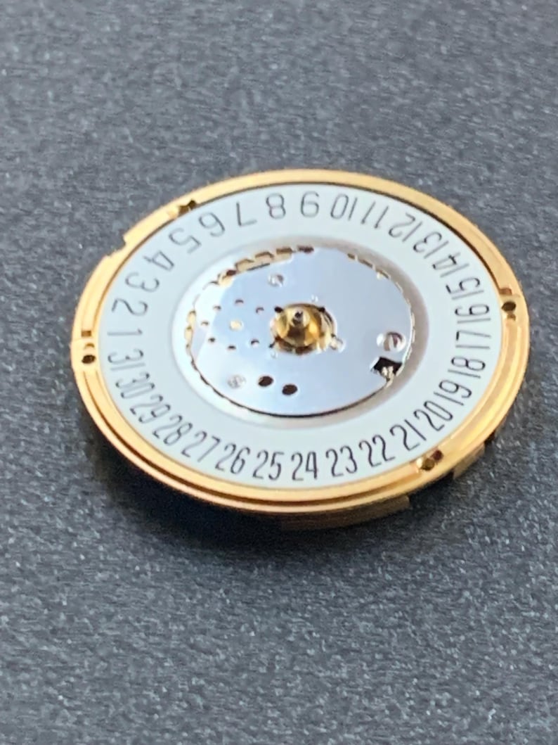 May include: A gold-toned watch movement with a white dial featuring black numerals from 1 to 31, indicating the days of the month. The central mechanism has a small gold-colored post. The watch component is circular.