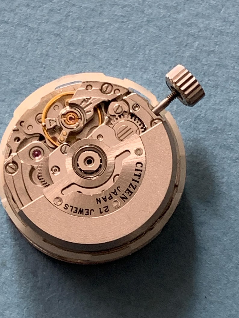 May include: Close-up of a Citizen watch movement with intricate gears, jewels, and a winding stem. The watch mechanism is silver-toned with the text "CITIZEN JAPAN 21 JEWELS" visible. The background is a solid blue.
