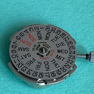 May include: A silver watch movement with a day and date dial. The dial displays the days of the week in English and the numbers 1-31. The text "334EG" is visible. The movement has a small crown and is set against a teal background.