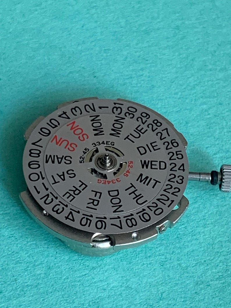 May include: A silver watch movement with a day and date dial. The dial displays the days of the week in English and the numbers 1-31. The text "334EG" is visible. The movement has a small crown and is set against a teal background.
